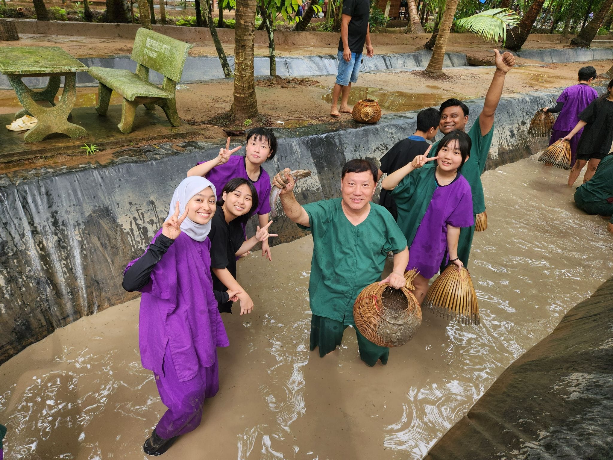 Group of people in purple and green outfits standing in muddy water, holding bamboo fish traps and posing cheerfully.