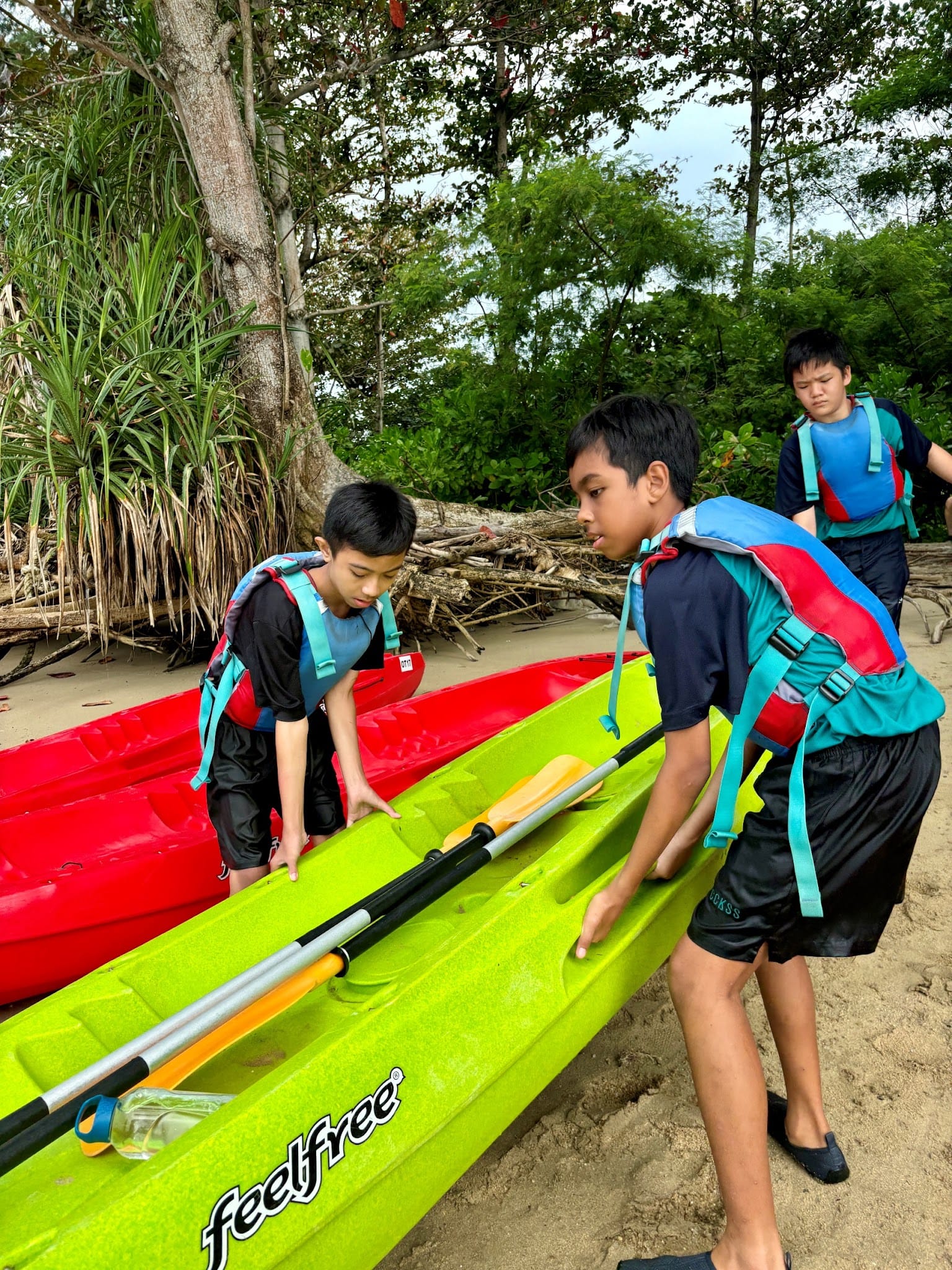 Three boys in life jackets handle a green Feelfree kayak with paddles on a sandy beach near trees.