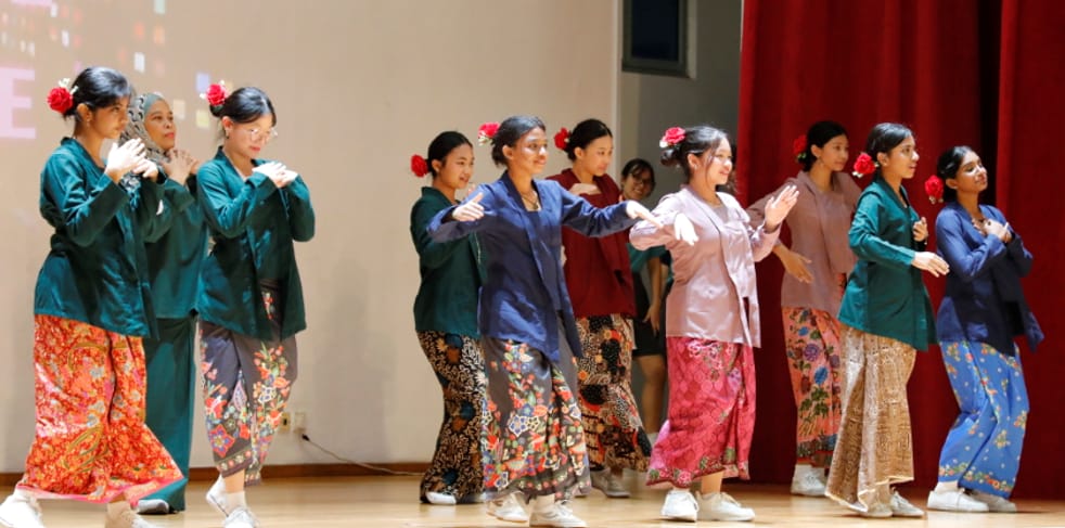 A group of women in colorful traditional batik skirts and red flowers in their hair perform a cultural dance on stage.