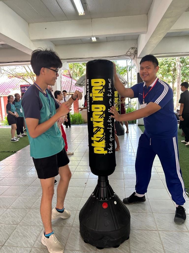 Two young men stand on either side of a black freestanding PKboxing Ring punching bag indoors.