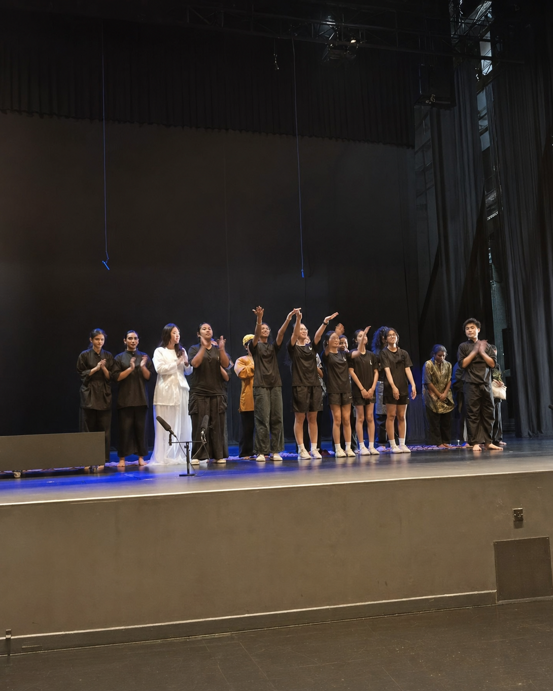 A group of performers bow and clap on a darkened stage after a show, most dressed in black.