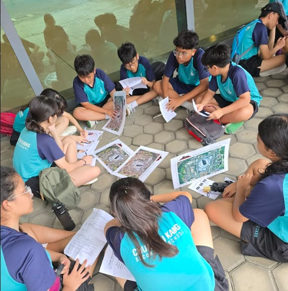 Students in teal uniforms sit in a circle on paved ground, reviewing printed maps and papers together outdoors.