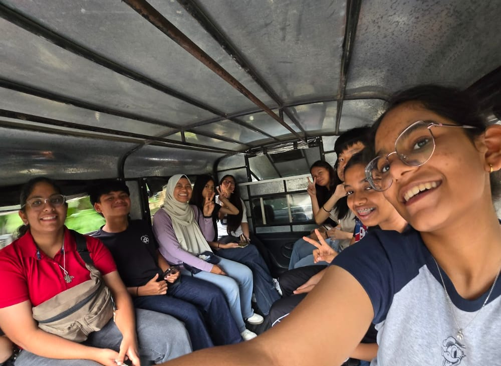 A group of smiling young people sitting inside a jeepney, several making peace signs, as one takes a selfie.