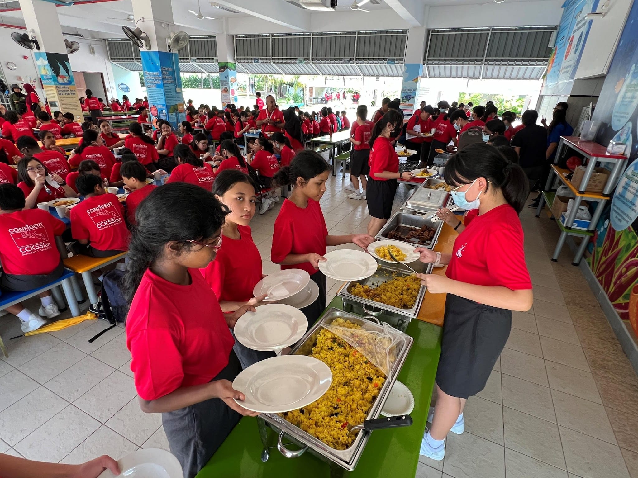 Students in red shirts hold plates in a cafeteria buffet line with trays of rice and other food.