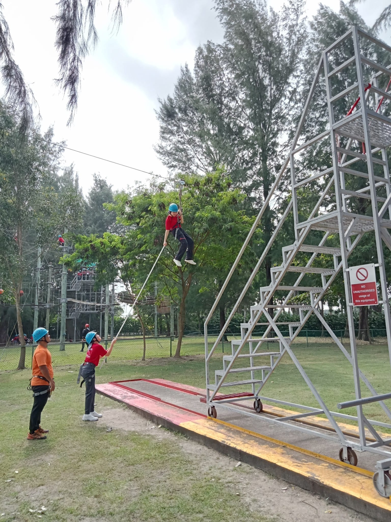 Person in blue helmet zip-lining from a tall metal tower while two helmeted instructors hold a rope below.