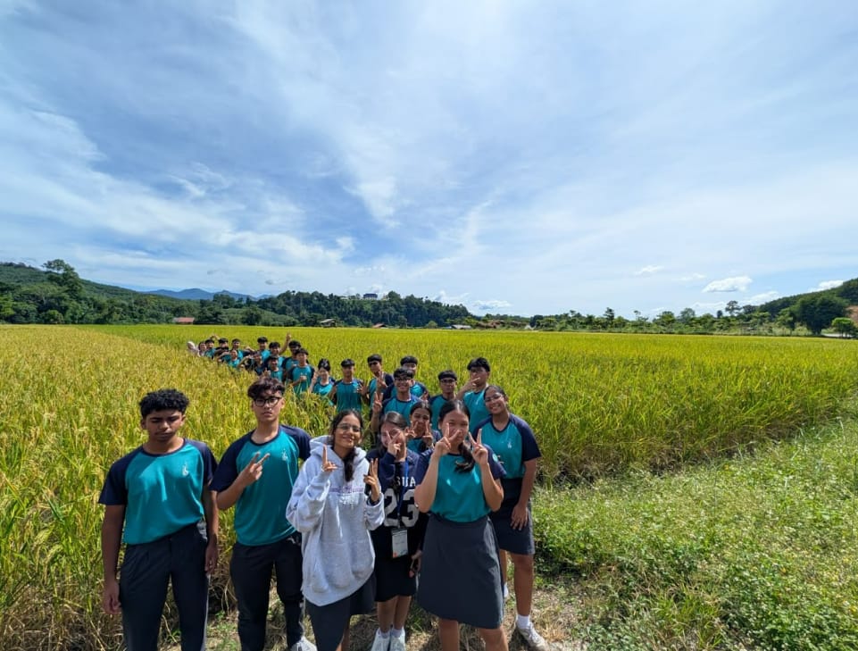 A group of students in teal uniforms pose with peace signs beside a golden rice field under a cloudy sky.