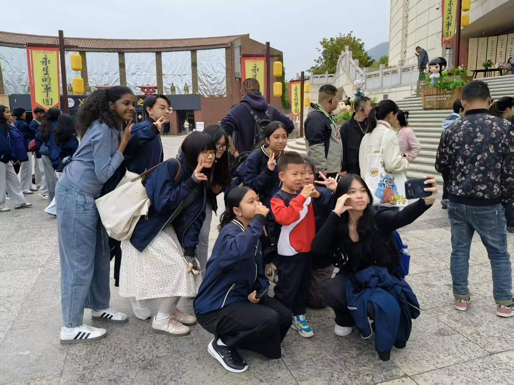 A group of children and teens pose for a selfie outdoors near a Chinese building with red banners and lanterns.