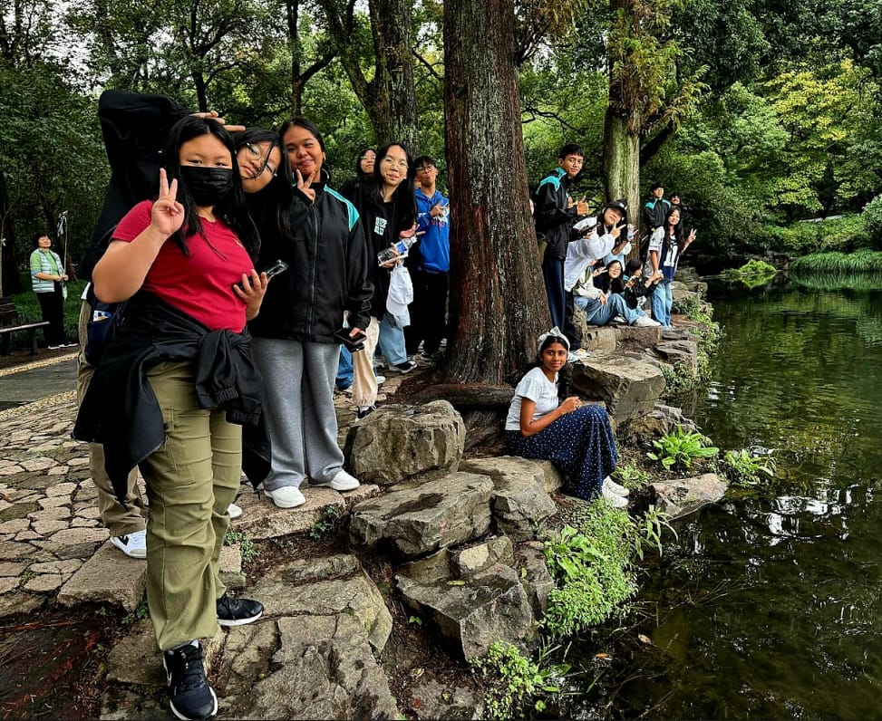 A group of teenagers posing with peace signs beside a pond on rocky steps in a lush green park.