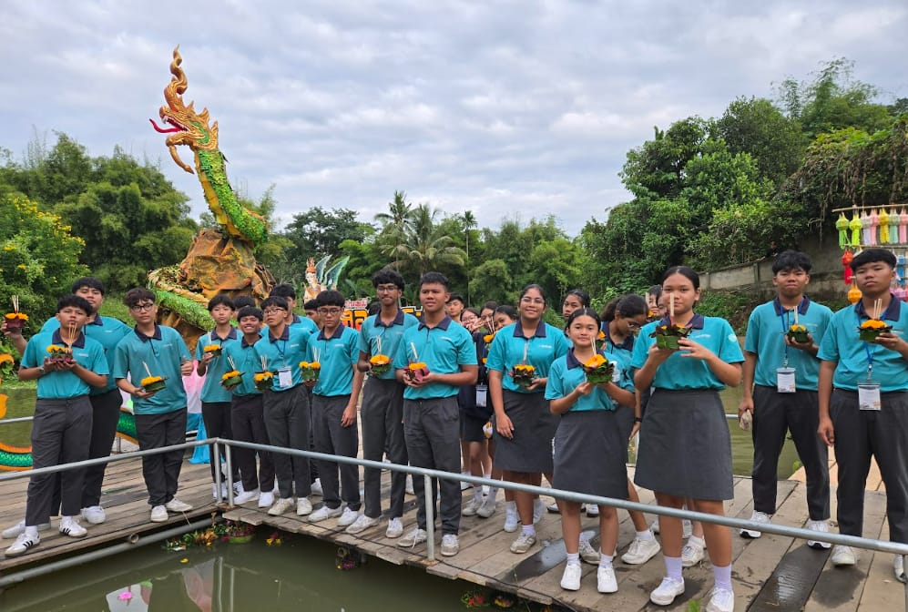 Students in teal uniforms hold flower floats on a wooden bridge near a decorative dragon statue by a river.