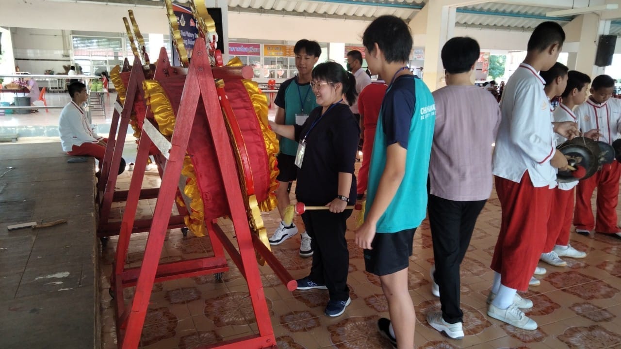 A girl strikes a large red and gold traditional drum on a stand while students in red pants play gongs nearby.