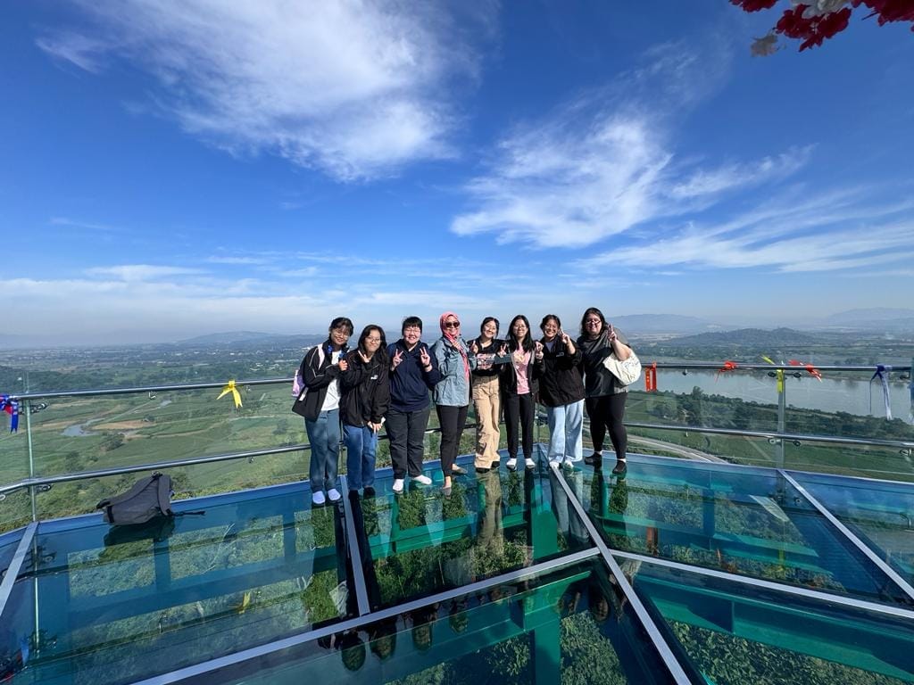Eight people pose on a glass-floor sky deck overlooking a green valley and river under a blue sky.