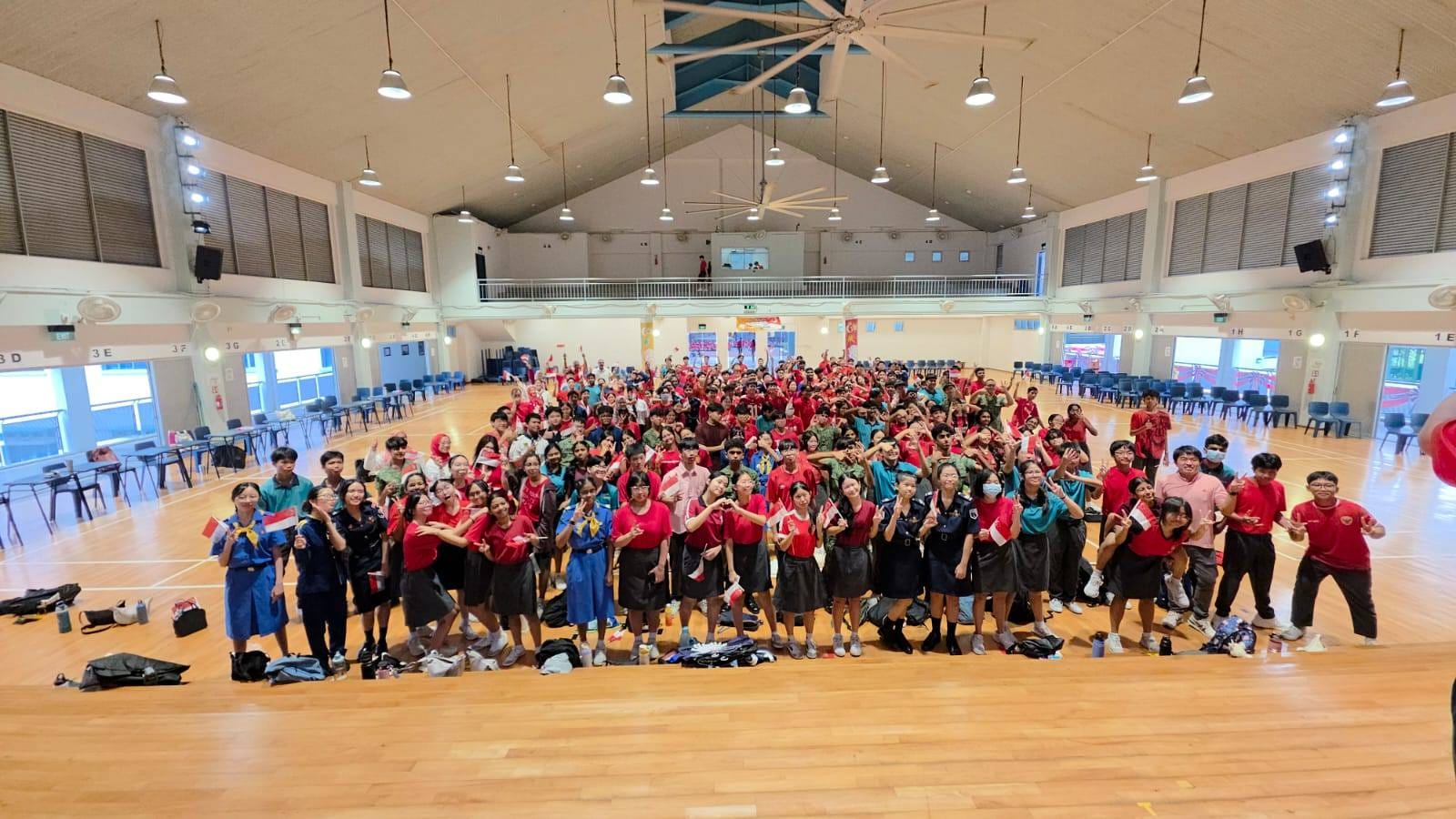 Large group of students in red uniforms posing together in a school hall, some holding small Singapore flags.