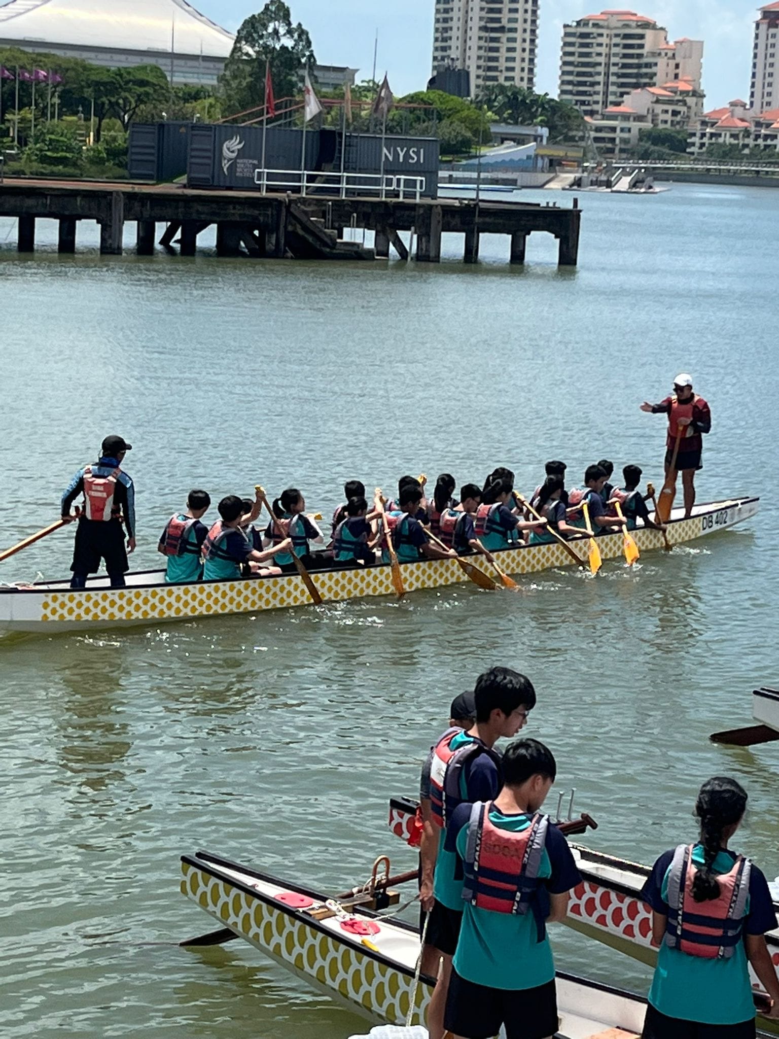 A team of young paddlers in life vests rows a dragon boat on a river near a dock with buildings behind.