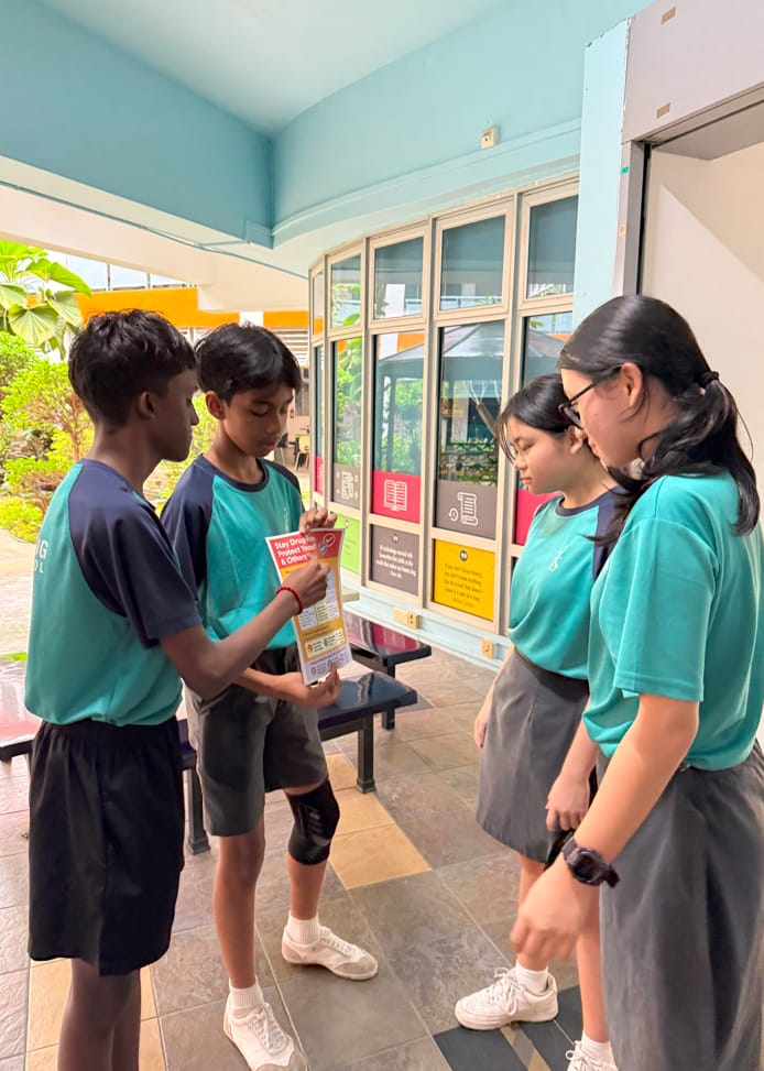 Four students in teal uniforms stand in a school corridor, reading a pamphlet about staying drug-free.
