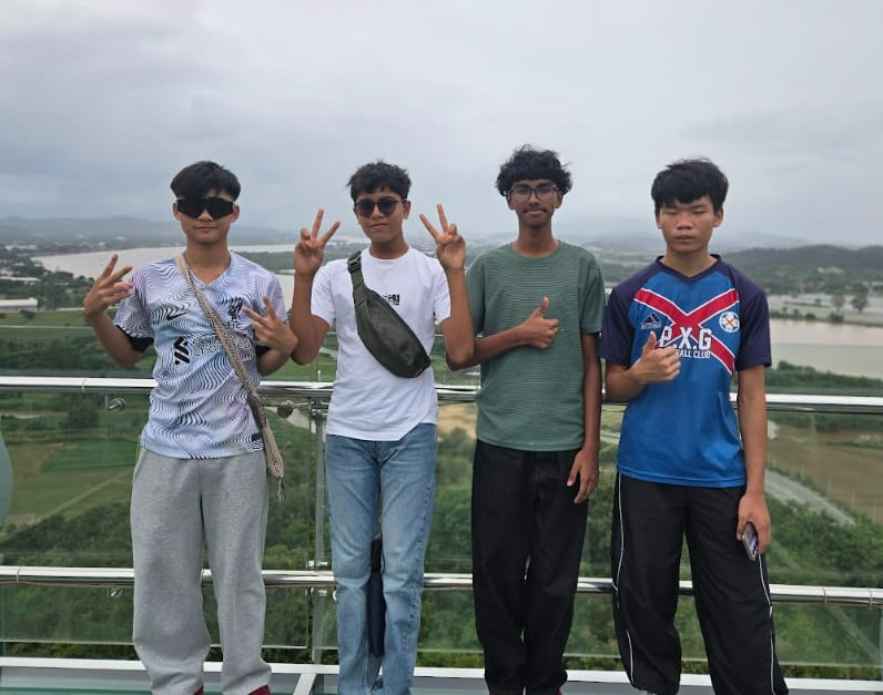 Four teenage boys pose on an elevated platform with peace signs, a river and cloudy sky visible behind them.