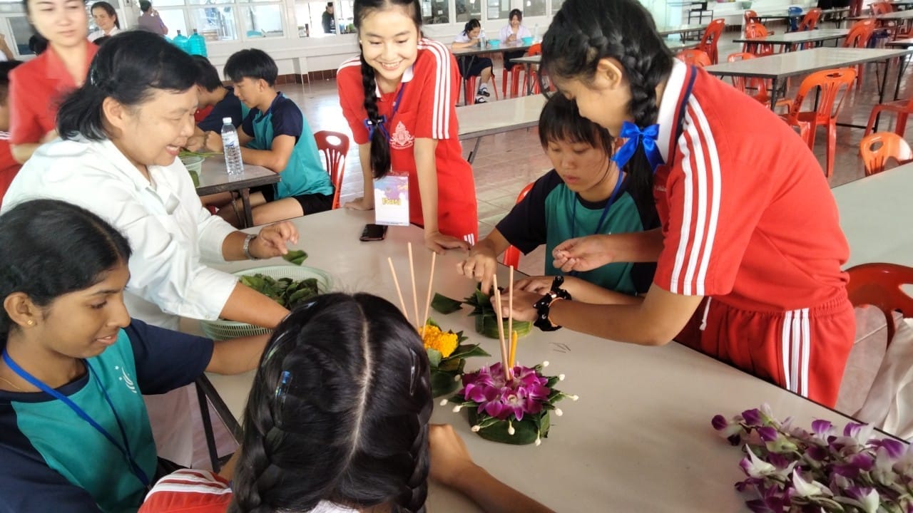 Several young people and an adult gather around a table assembling a krathong float decorated with flowers and incense sticks.