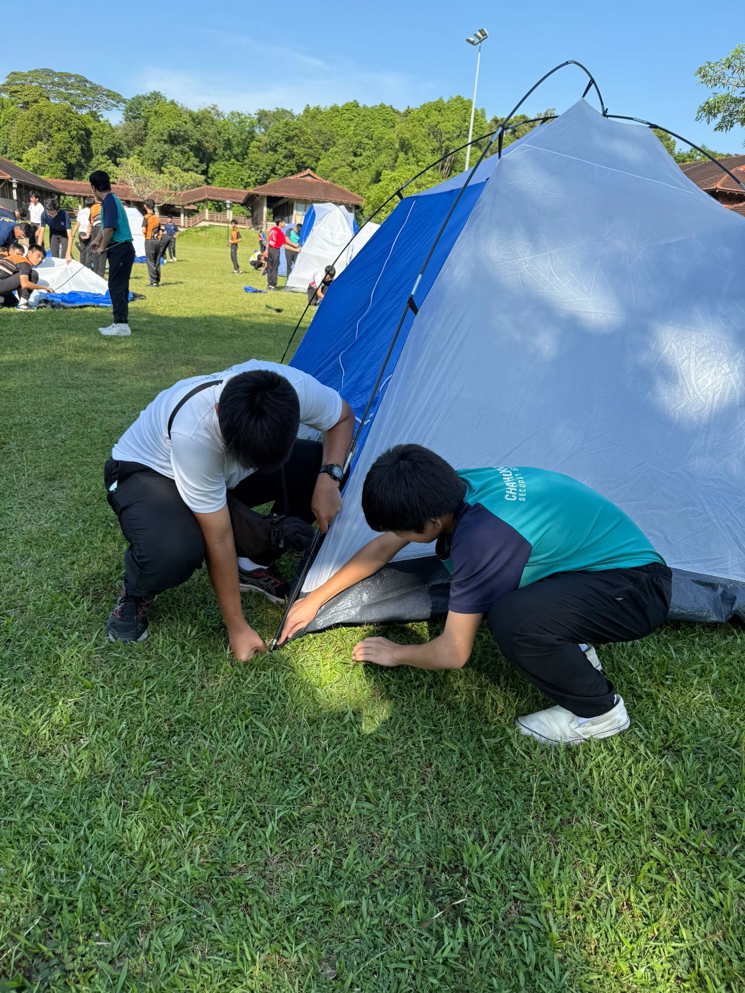 Two boys kneeling on grass securing the base of a blue and grey tent at an outdoor campsite.