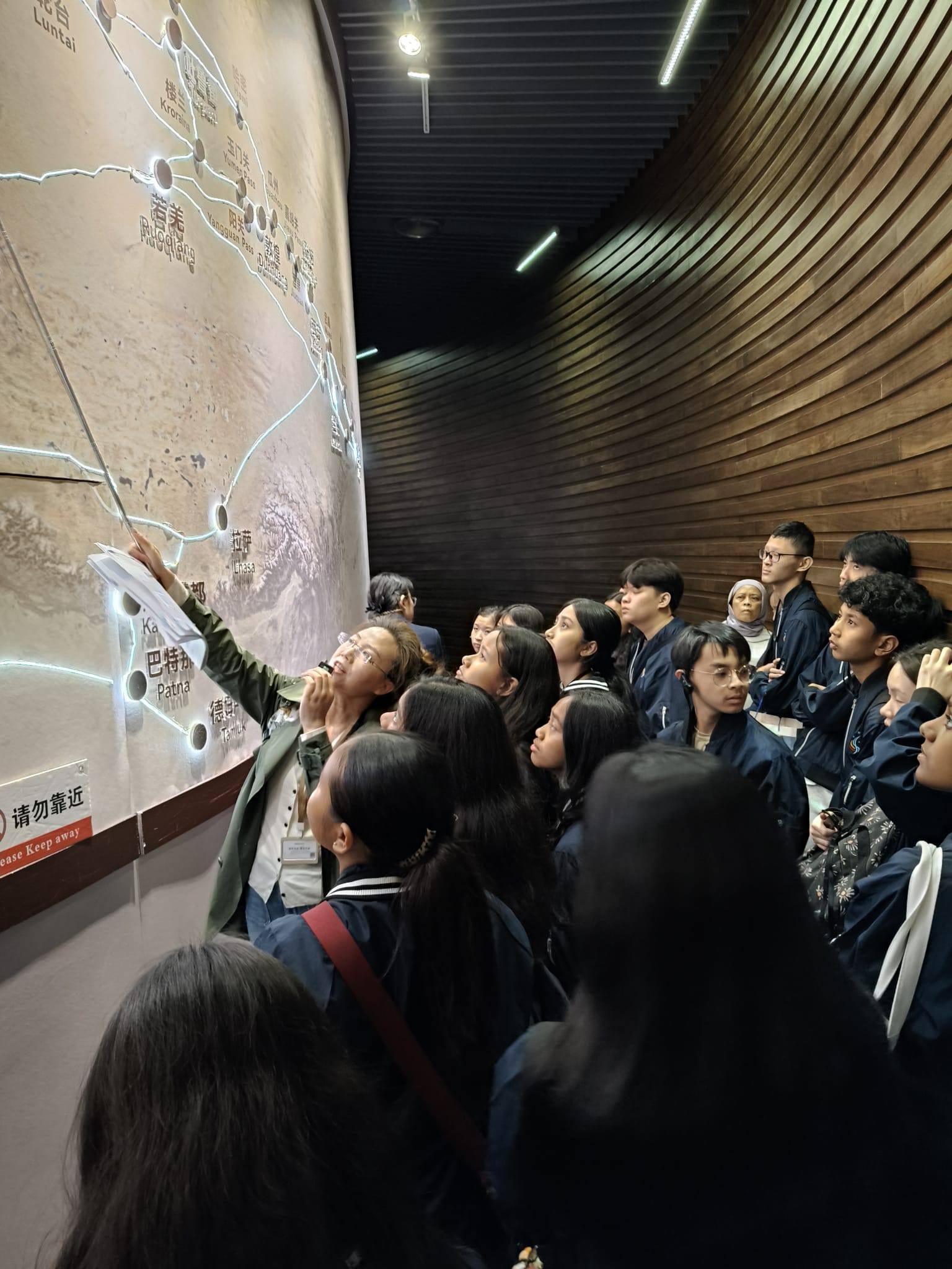 A guide points to a large illuminated wall map with Chinese text while a group of students looks on attentively.