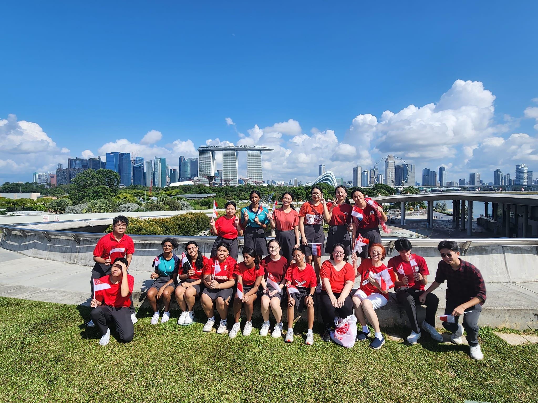 Group of people in red shirts holding small Singapore flags pose outdoors with Marina Bay Sands skyline behind them.
