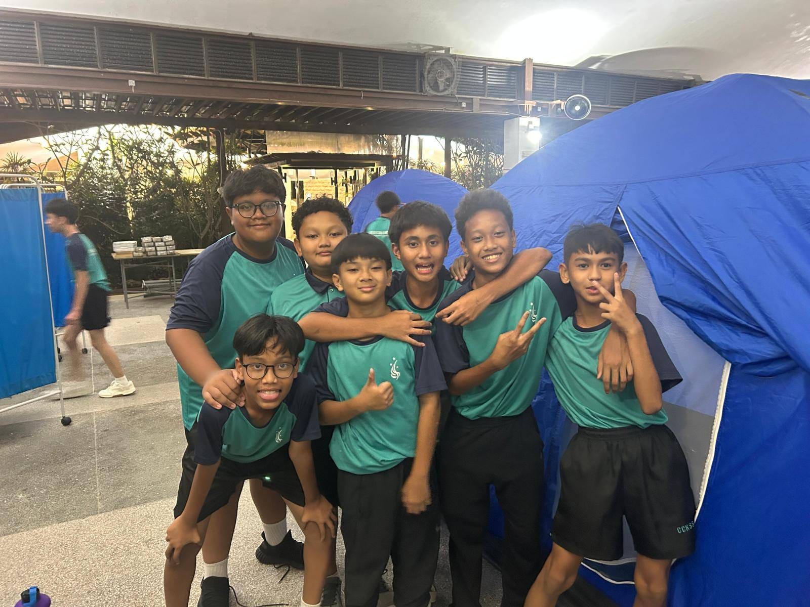 Six boys in matching teal uniforms pose together smiling near blue camping tents in an indoor venue.