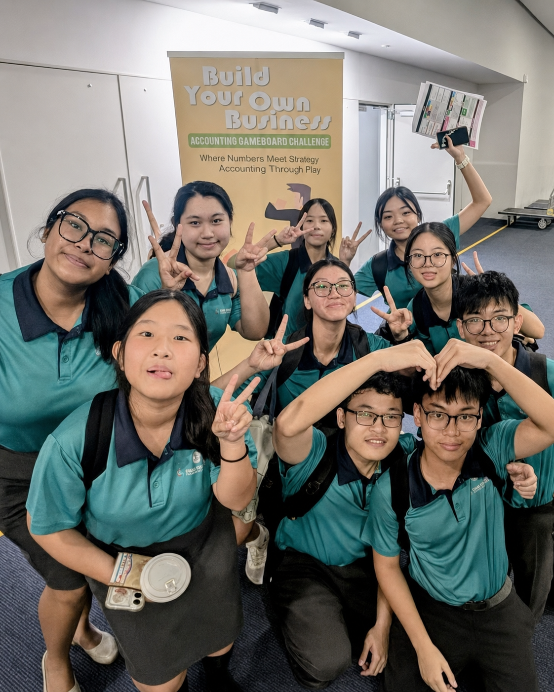 Ten students in teal uniforms pose with peace signs before a Build Your Own Business Accounting Gameboard Challenge banner.
