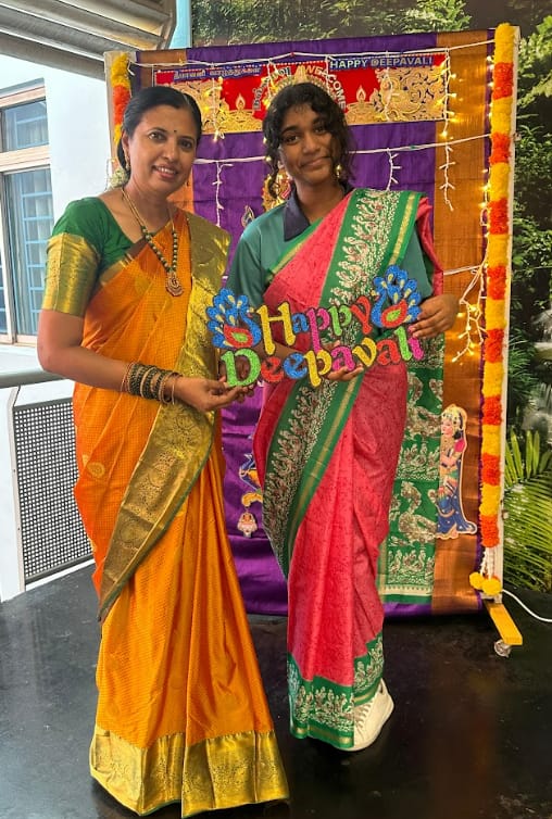 Two women in colorful sarees hold a Happy Deepavali sign in front of a decorated Diwali backdrop.