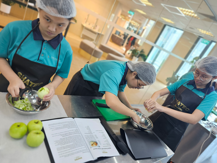 Three students in teal uniforms, black aprons, and hairnets prepare green apples at a kitchen counter with an open recipe book.
