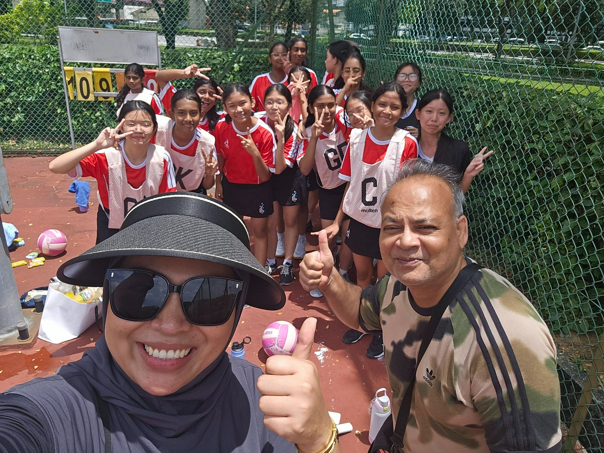 Two adults give thumbs up in a selfie with a group of girls in red netball uniforms on an outdoor court.