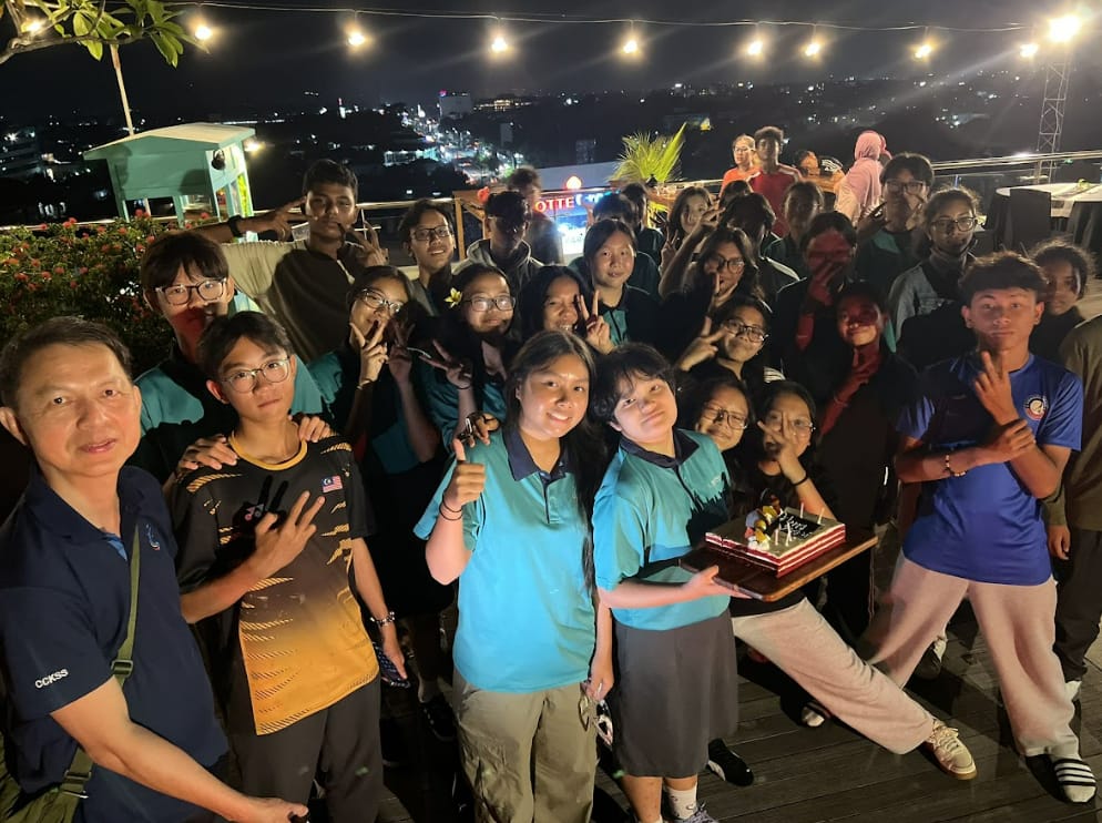 Large group of young people posing at night on a rooftop, one person holding a birthday cake with city lights behind.
