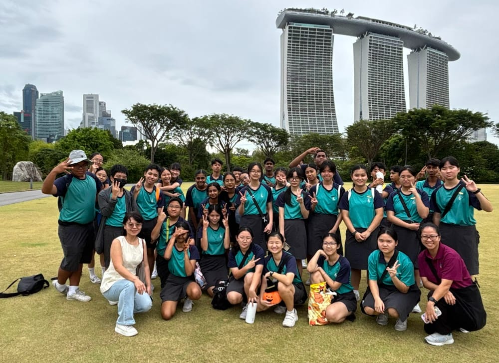 A large group of students in teal uniforms pose on grass with Marina Bay Sands towers in the background.