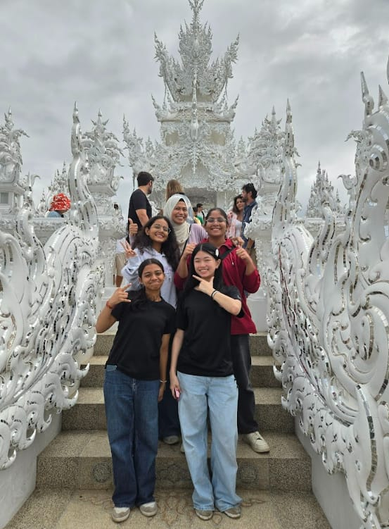 Four young women pose on steps in front of the ornate white Wat Rong Khun temple in Thailand.
