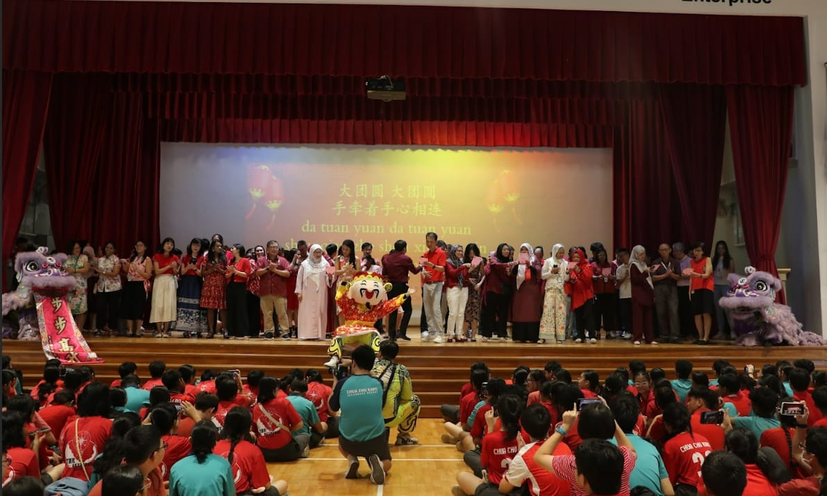 A large group on stage celebrates Chinese New Year with lion dancers, a mascot, and Chinese lyrics projected behind them.