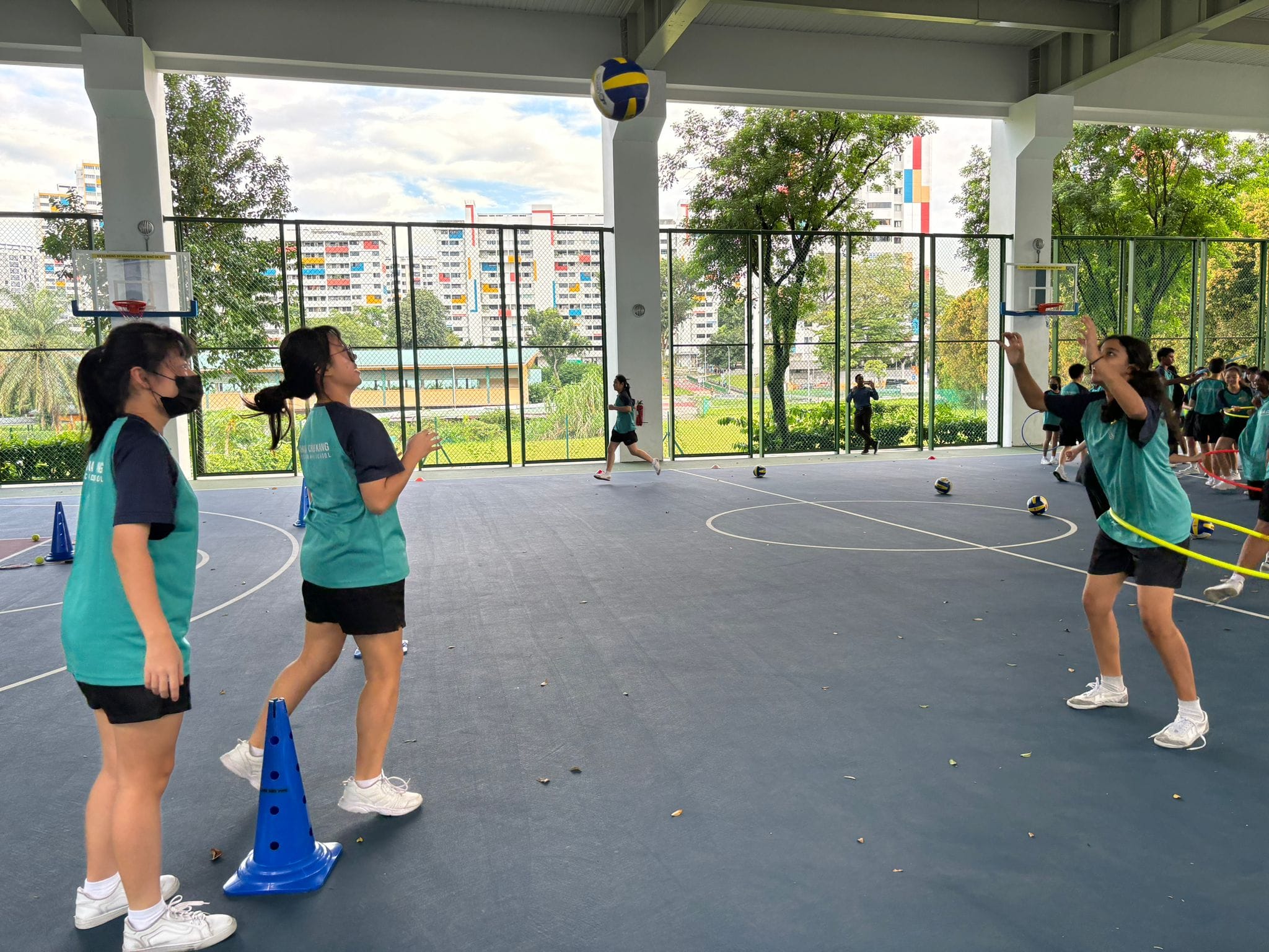 Students in teal shirts practice volleyball on an outdoor covered court with blue cones and city buildings in the background.