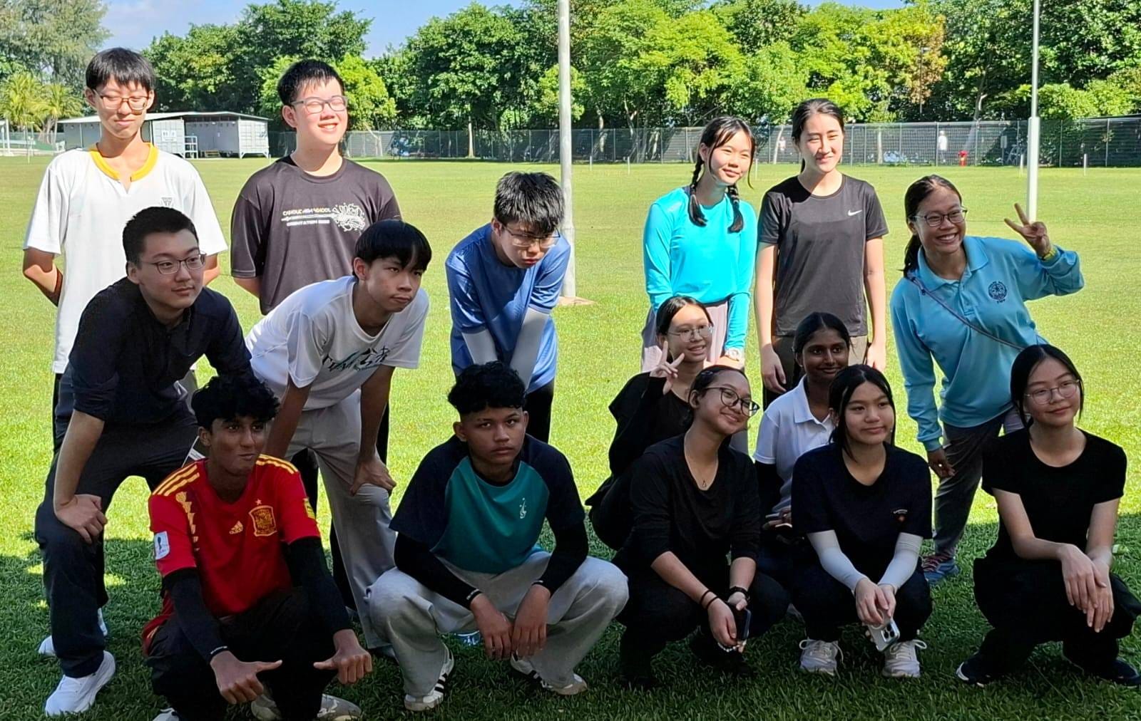 A group of about 15 teenagers posing together on a green sports field surrounded by trees.