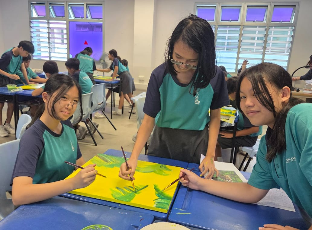 Three students in teal uniforms paint green plants on yellow canvas during an art class.