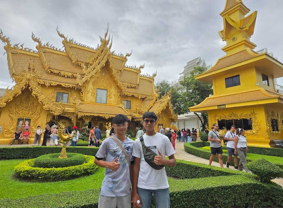 Two young men pose in front of an ornate golden Thai temple with manicured hedges and crowds of visitors.