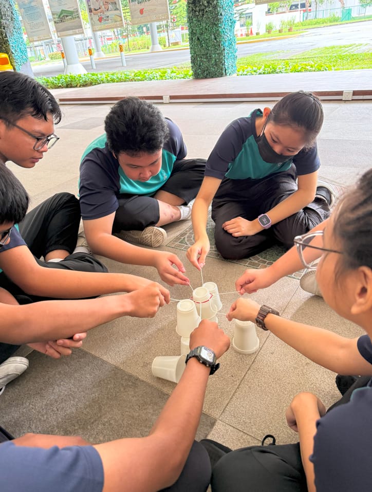 Five students in uniforms sit on the floor using strings attached to a rubber band to stack paper cups together.