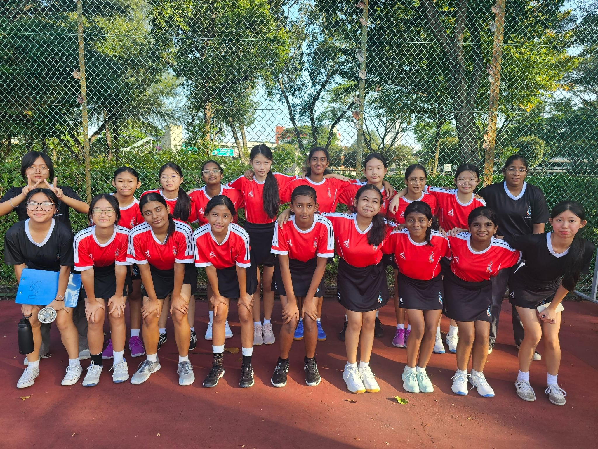 A group of young girls in red and black sports uniforms posing together on an outdoor court near a chain-link fence.
