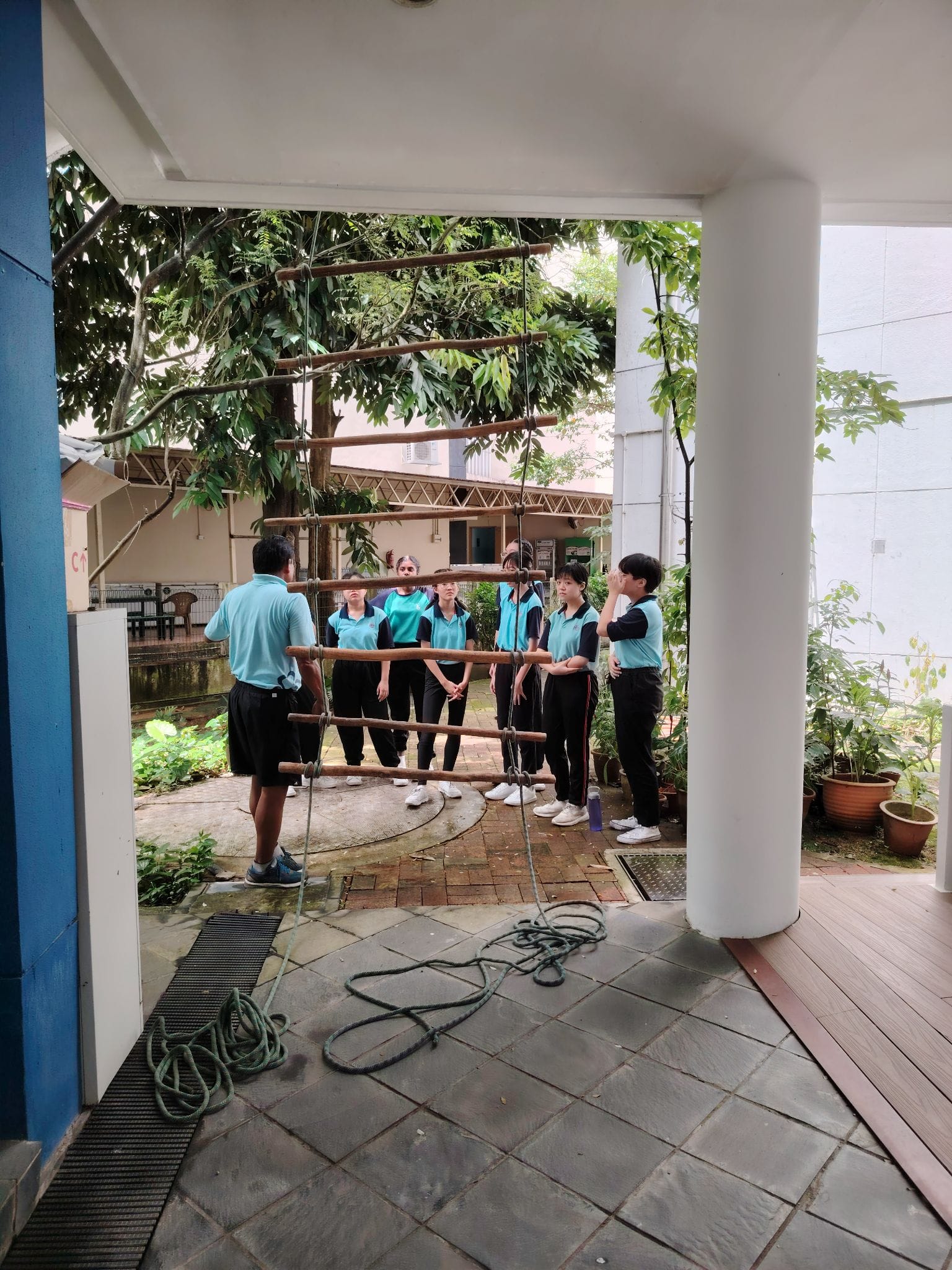 Students in teal shirts and black pants stand around a suspended rope ladder during an outdoor activity at school.