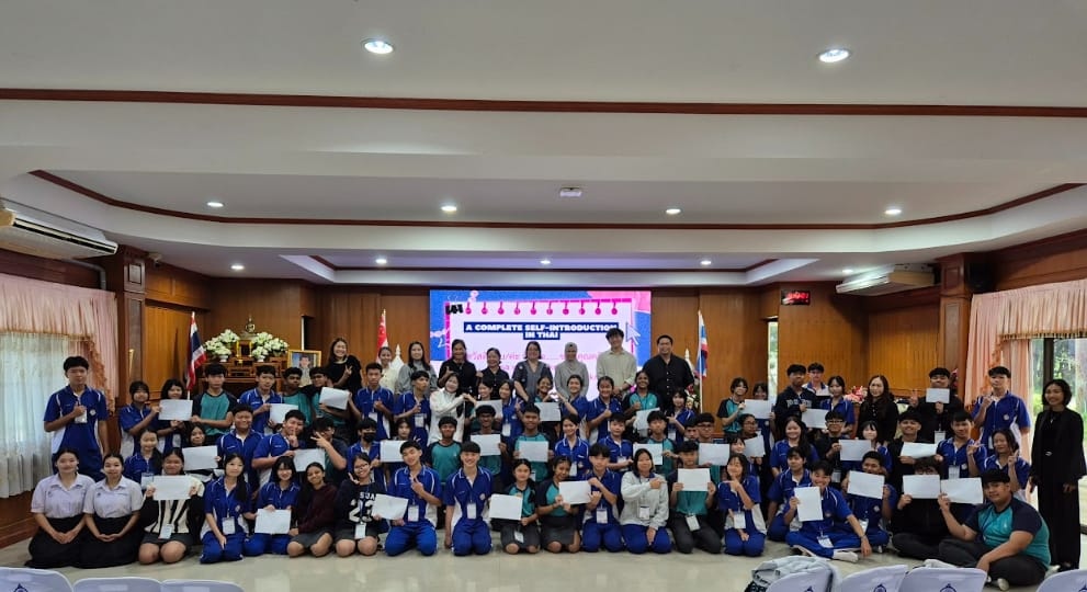 Large group of students in blue uniforms holding certificates in a hall with a screen reading "A Complete Self-Introduction in Thai."