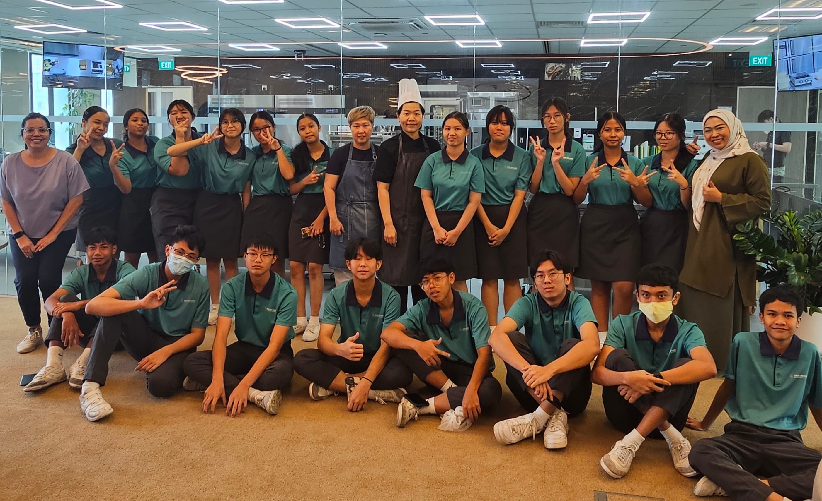 A large group of students in teal uniforms poses with two instructors wearing chef aprons indoors.