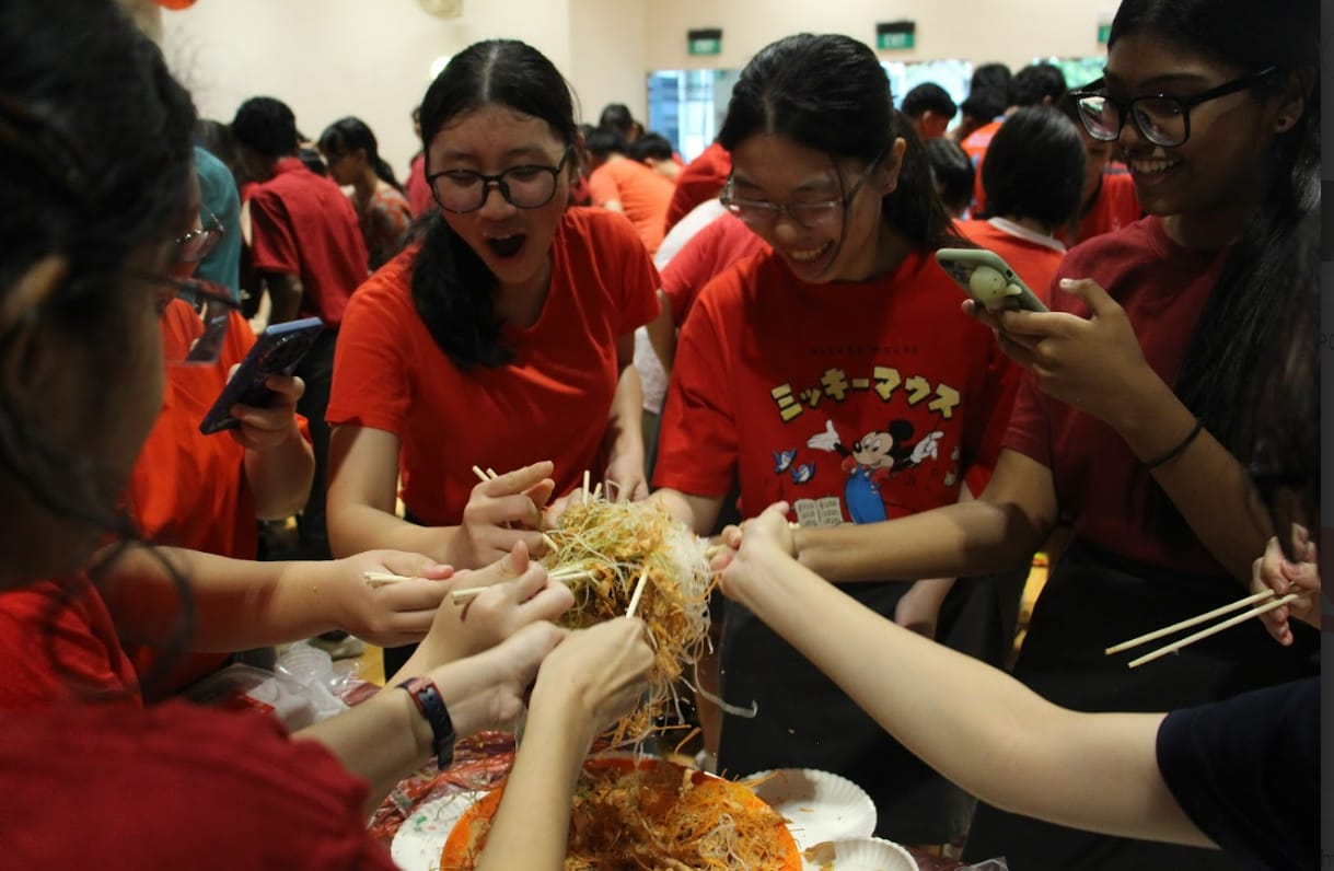 A group of young people in red shirts joyfully tossing yusheng with chopsticks at a festive gathering.