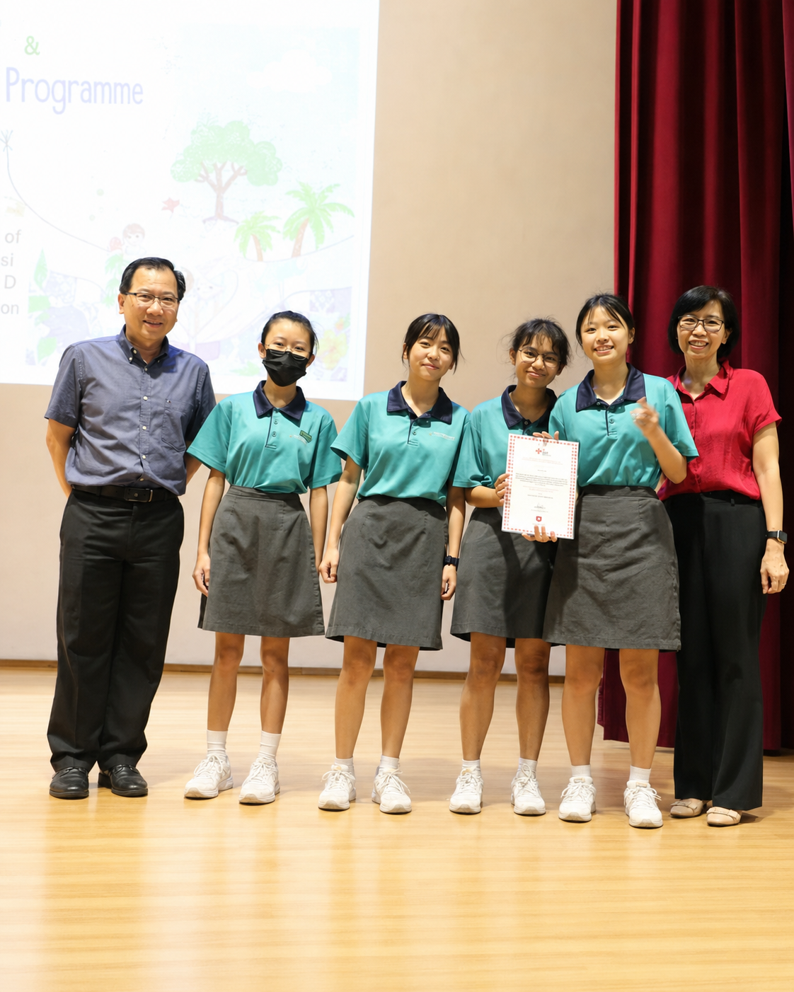 Six people pose together on a stage, with three students in teal uniforms holding a certificate.
