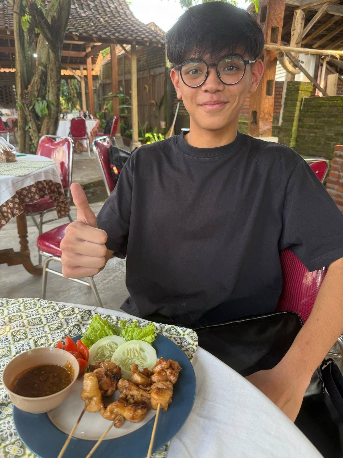 Young person with glasses giving thumbs up at a restaurant table with a plate of satay skewers and dipping sauce.