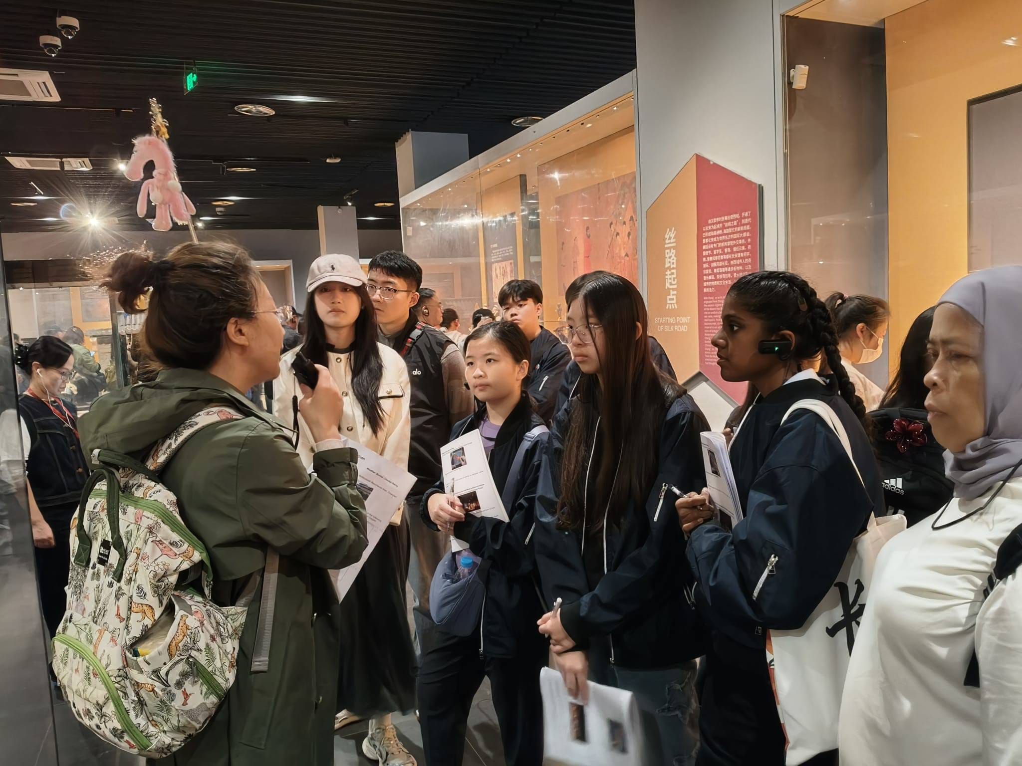 A guide speaks to a diverse group of visitors holding pamphlets inside a Silk Road museum exhibit.