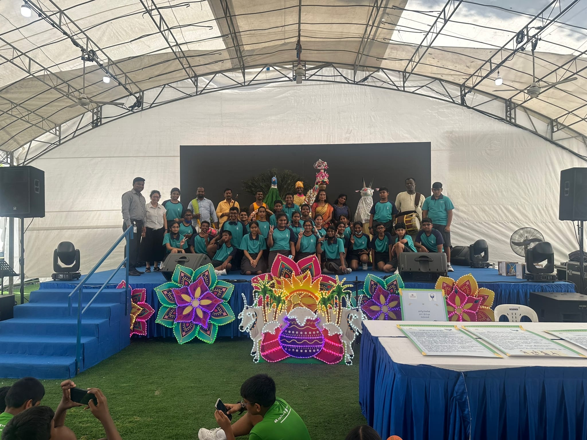 A large group of students in teal uniforms pose on a decorated stage with colorful floral light displays at a cultural event.