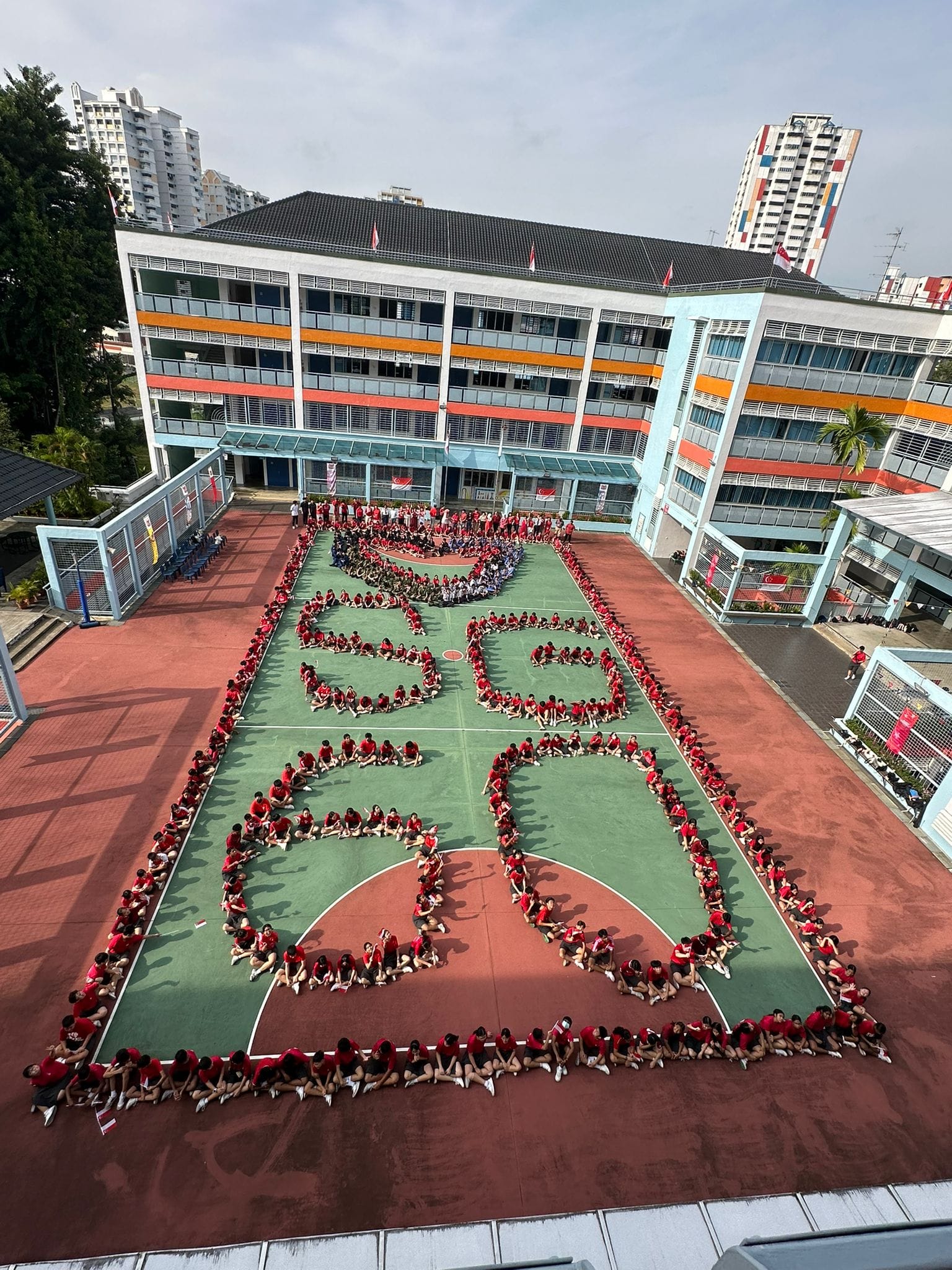 Hundreds of students in red shirts form the number 60 on a school basketball court, viewed from above.