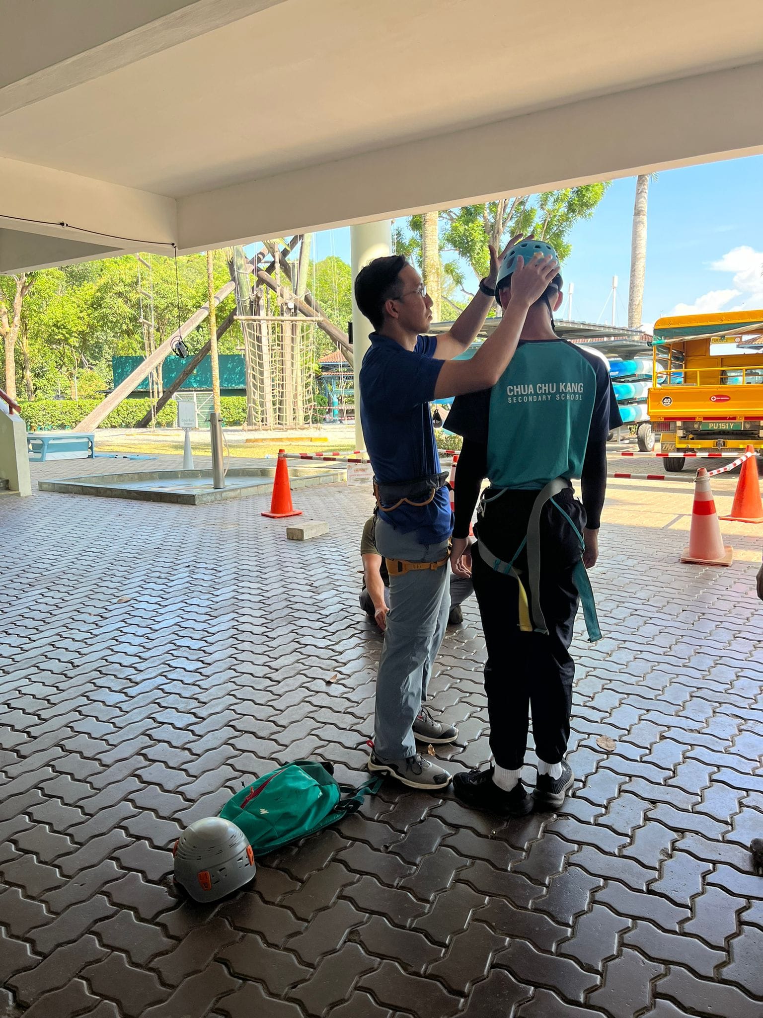 An instructor adjusts a blue helmet on a student wearing a harness and Chua Chu Kang Secondary School shirt outdoors.