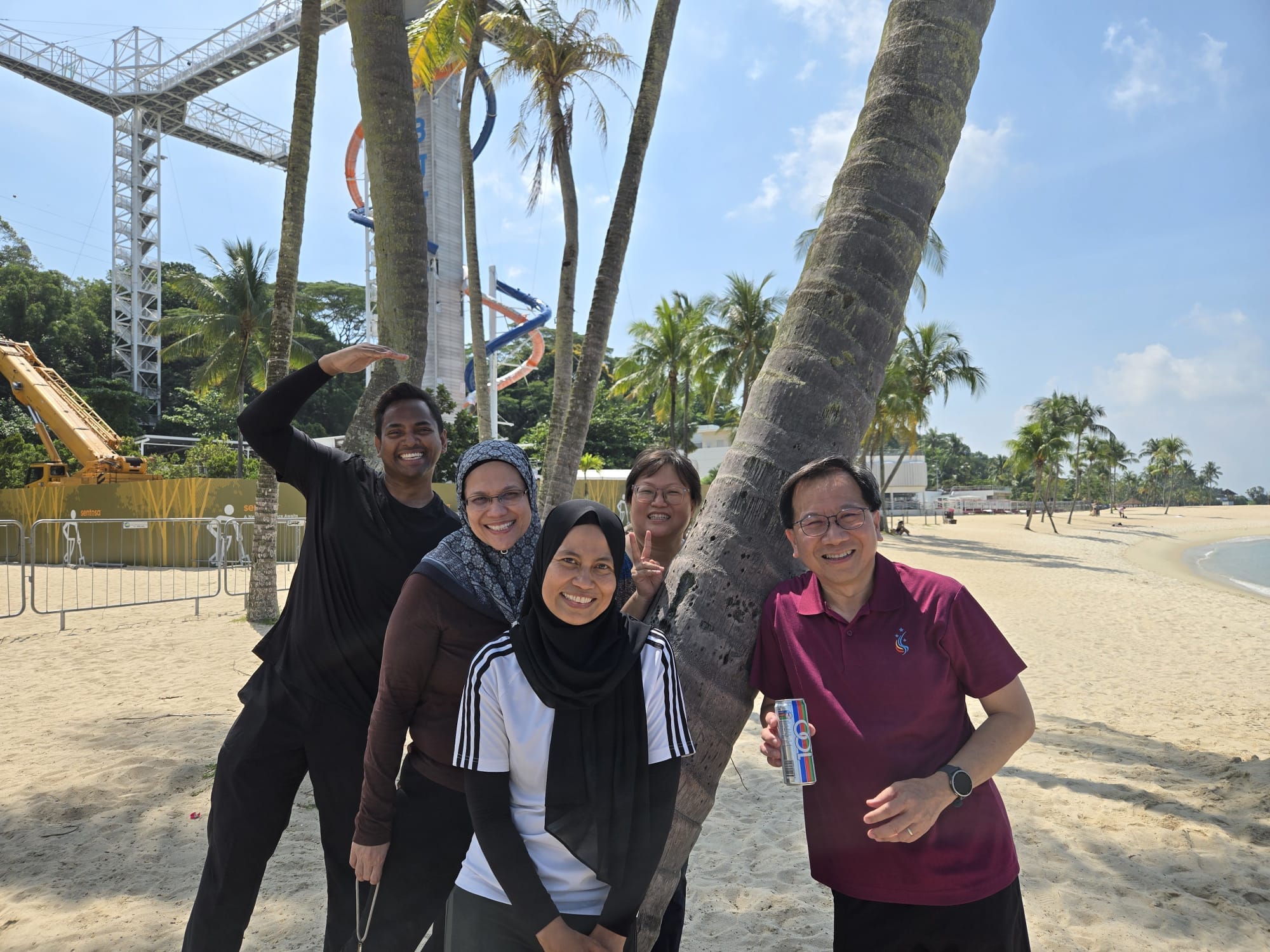 Five smiling people pose on a sandy beach near palm trees, with colorful water slides and a tall tower in the background.