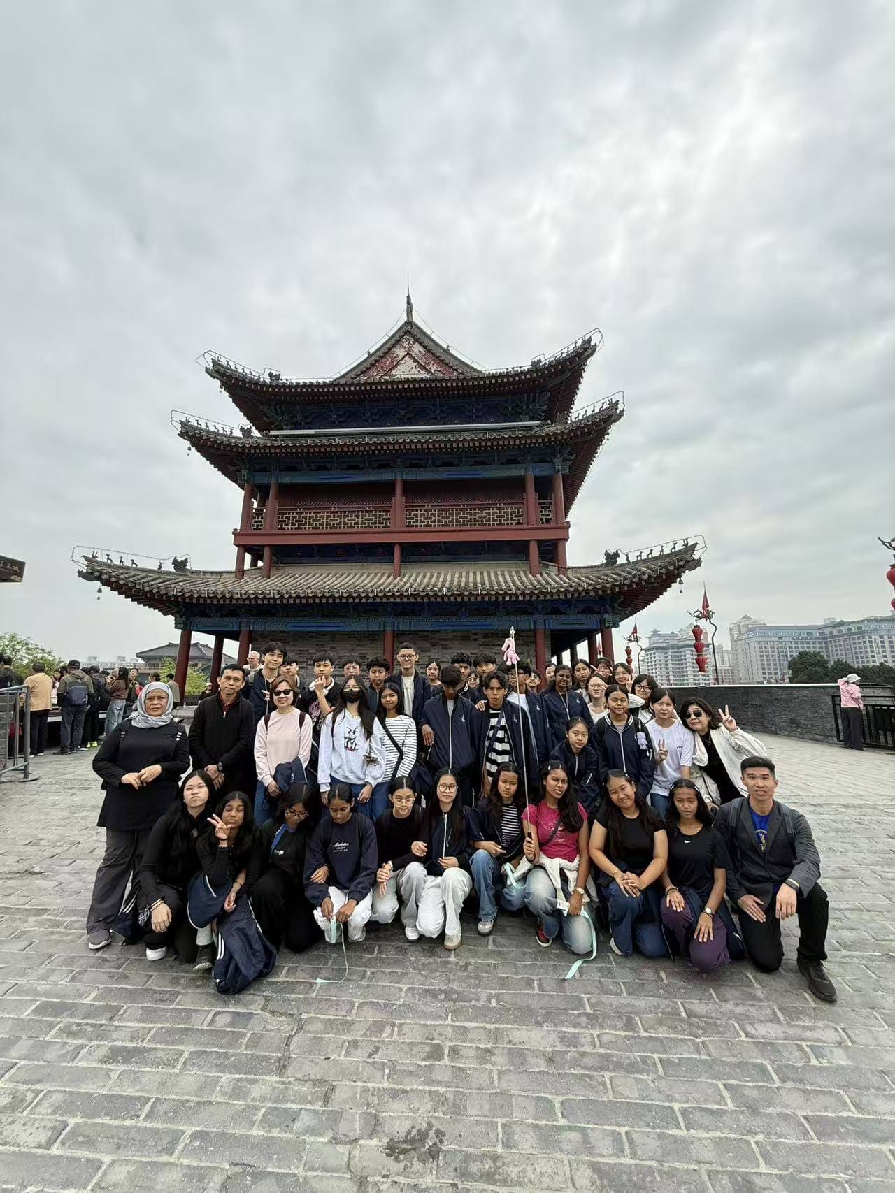 A large group of young tourists poses in front of a multi-tiered traditional Chinese pagoda under a cloudy sky.